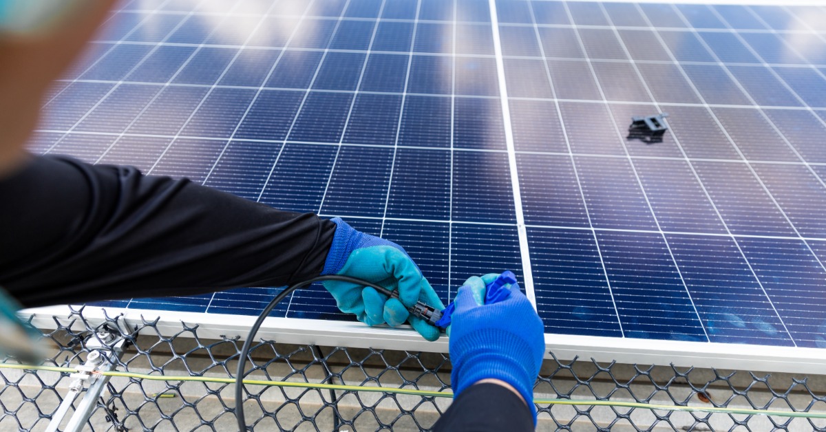 A Look at the Key Components of a Modern PV System 1 A close-up of the hands of a worker wearing blue gloves, holding a black cable attached to the back of a solar panel.
