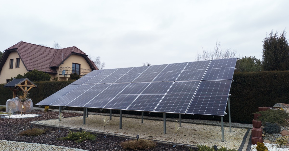 How To Optimize Solar Panel Tilt for Summer Sun Exposure 1 Two long rows of solar panels aligned next to each other, placed on a ground structure next to a two-story home.