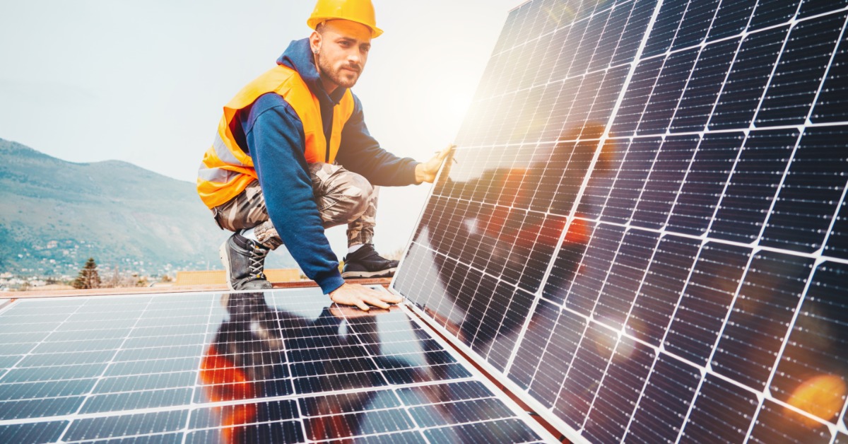 A worker wearing a vest and a helmet, touching a solar panel flat on a roof while holding another panel at an angle.
