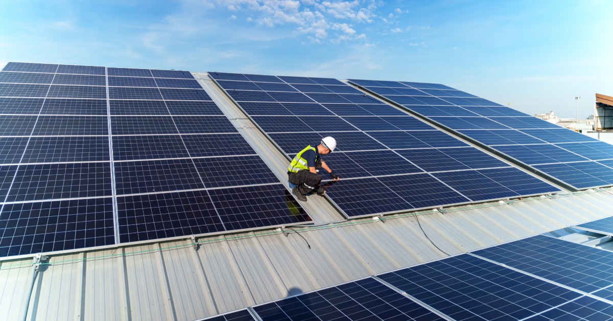A worker wearing a safety vest and a helmet fixing a solar panel on the corner of a section with interconnected panels.