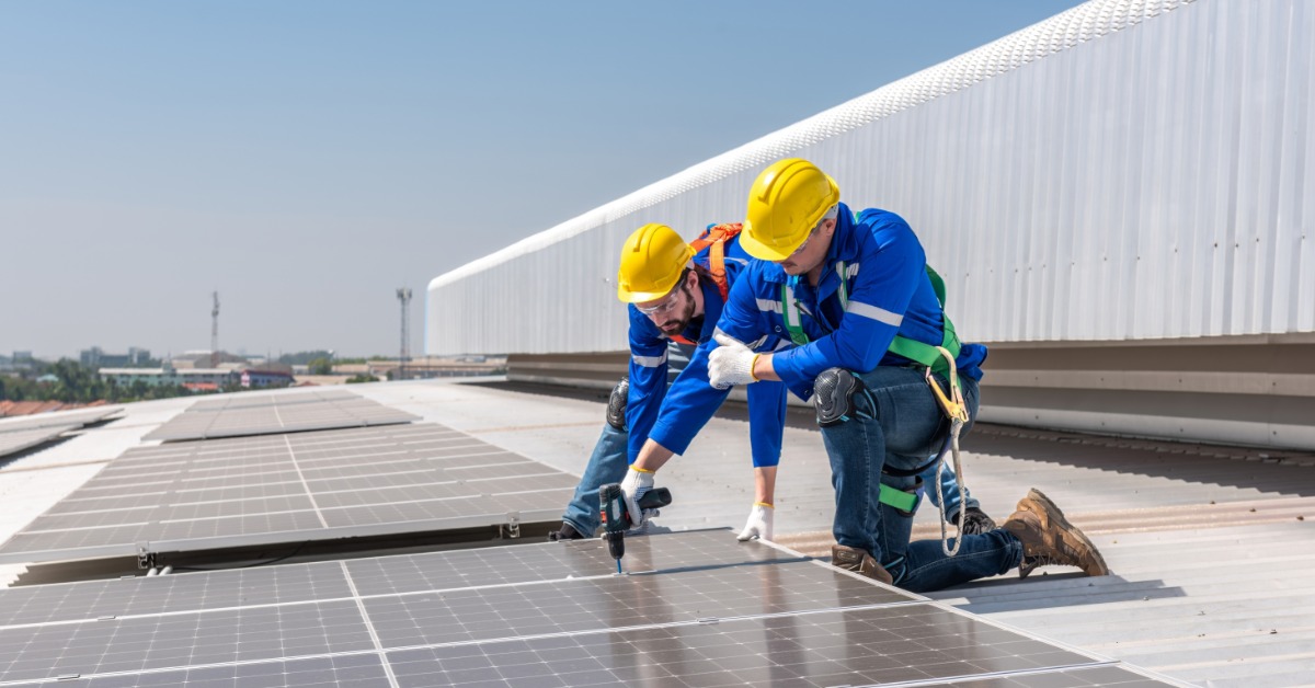 Two workers wearing yellow helmets drilling in place a solar panel on a commercial roof next to other panels.