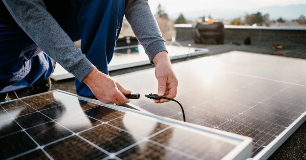 Megapack vs. Powerwall: Core Differences 1 A close-up of two hands plugging two black cables together over multiple large solar panels placed on a roof.