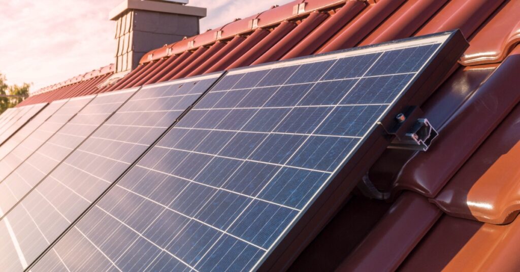 A close-up of a row of solar panels on a residential roof, over brown, clay shingles, with the sun in the background.