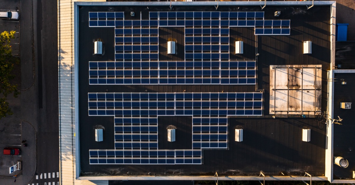 An aerial view of a large building with multiple rows of solar panels on the roof, surrounded by a large parking lot.