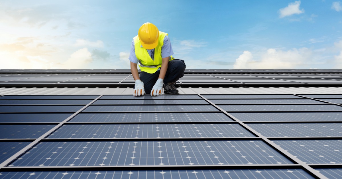 A worker wearing a yellow vest, helmet, and white gloves, touching a solar panel on a long row of panels.