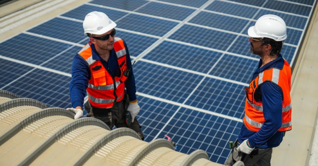 Two workers wearing safety vests and white helmets, standing on the roof of a building with multiple solar panels.
