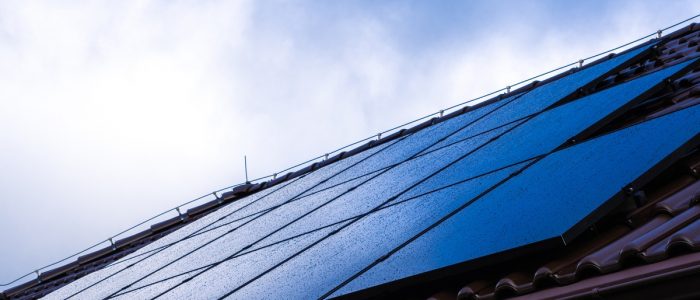 Multiple rows of solar panels on top of a roof with clay shingles, covered in water drops under a cloudy sky.