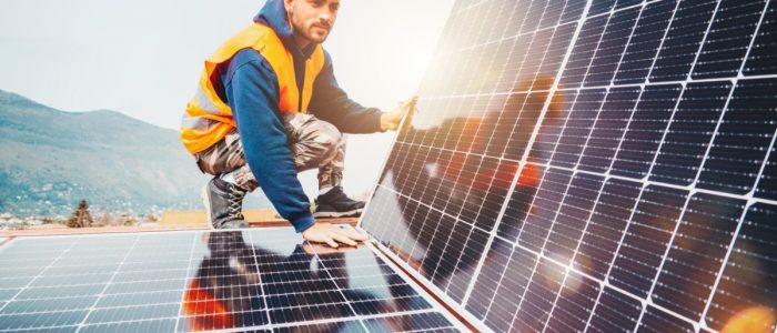 A worker wearing a vest and a helmet, touching a solar panel flat on a roof while holding another panel at an angle.