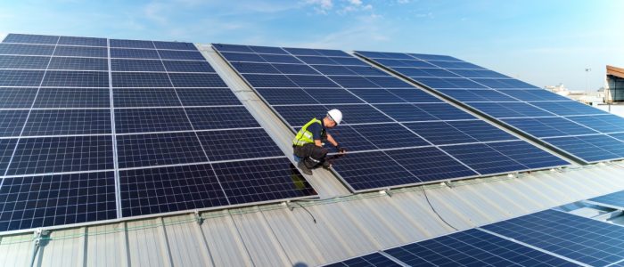 A worker wearing a safety vest and a helmet fixing a solar panel on the corner of a section with interconnected panels.