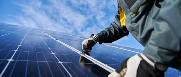 A close-up of a worker wearing a thick jacket and gloves touching the side of a solar panel from a long row of panels.