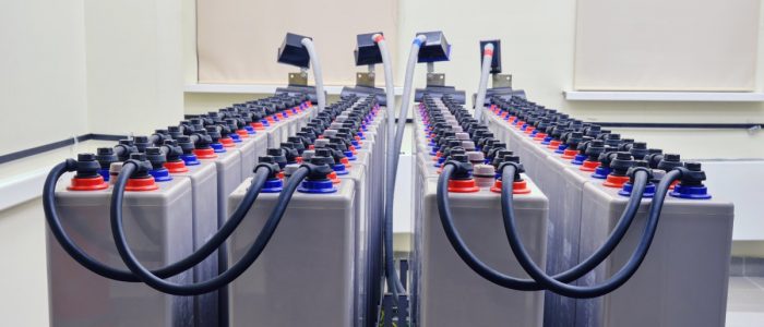 Multiple long gray batteries with interconnected cables placed next to each other inside a large white power room.