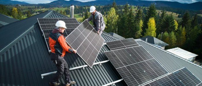 Two workers wearing white hard hats install a large solar panel on the roof of a home next to other panels.