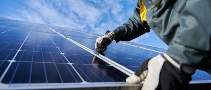 Close-up of a man wearing safety gloves and a jacket securing a large solar panel next to other panels on a roof.