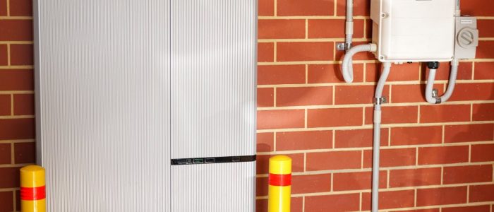 A large white home battery attached to a brick wall with a small transformer on the side attached to white tubes.