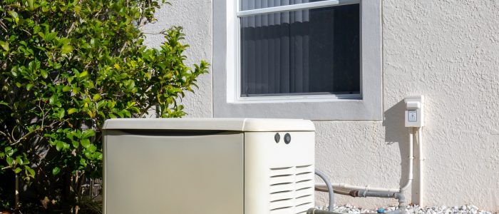 White residential power generator placed outdoors under a window, next to a bushy tree and lush green grass.