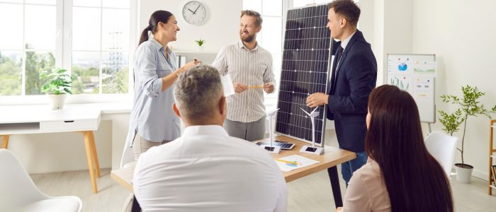 A group of people gathered in a meeting room, talking together. One man is holding a small solar panel on top of a desk.