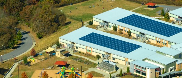 Aerial view of a school with a large parking lot and two buildings, each one with multiple solar panels on the roof.