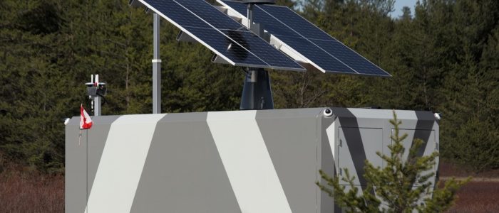 Large grey and white battery with solar panels attached to the top, placed on a plot of land surrounded by trees.