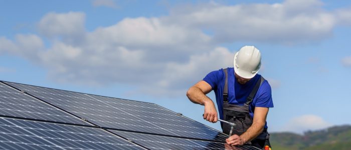 A worker wearing a blue T-shirt and white hard hat uses a wrench to tighten a solar panel installed on a roof.