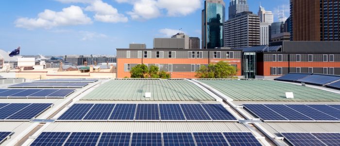 An aerial view of multiple solar panel rows on a large roof with multiple buildings in the background on a sunny day.