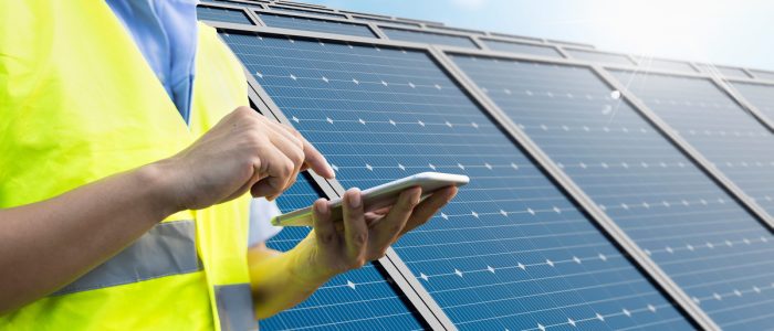 A close-up to the hands of a man wearing a yellow vest, holding a smartphone with solar panels in the background.