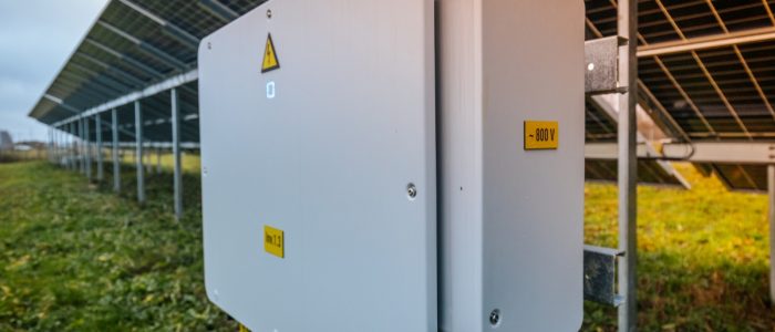 A close-up of a large inverter box placed under multiple rows of commercial solar panels in an open grass field.