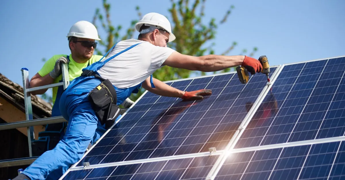 Everything You Need To Know About Solar Panels in 2025 1 Two workers wearing helmets and protective gloves standing on top of a roof installing multiple large solar panels.