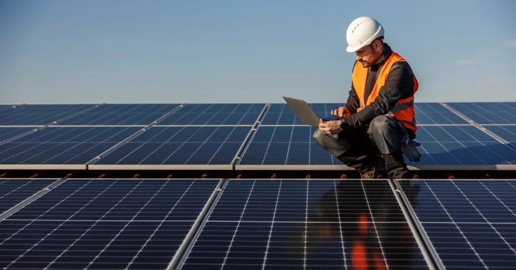 Man wearing an orange vest and a white helmet using a laptop while squatting on top of a roof covered with solar panels.