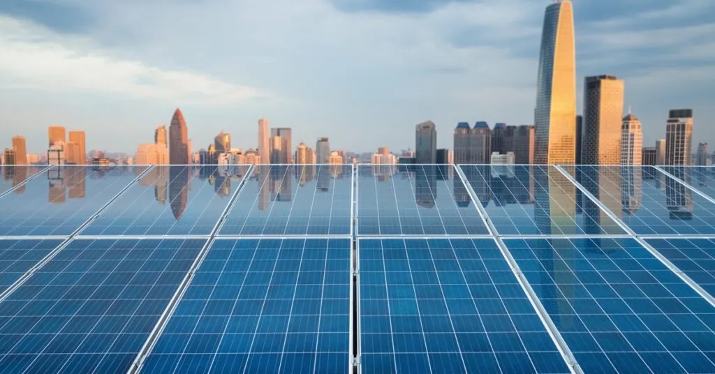 Multiple large solar panels in two rows with skyscrapers and buildings in the background on a cloudy day.