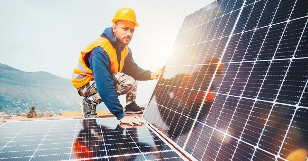 Why Hiring Solar Service Experts Matters 1 A worker wearing an orange hard hat and safety vest squats while installing a large solar panel on a roof.