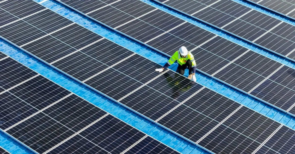 A worker wearing a bright yellow jacket and a white hard hat kneels next to a row of solar panels on a roof.