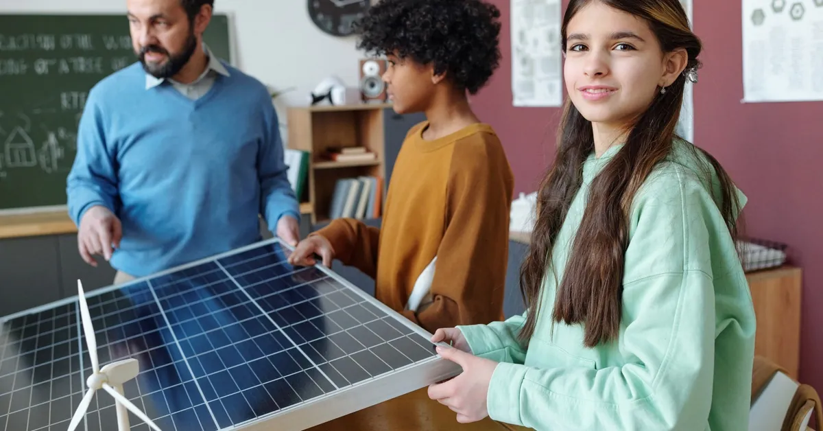 How Schools Are Benefiting From Commercial Solar Power 1 In a classroom setting, two young students and a professor are holding a small solar panel placed on a wooden desk.