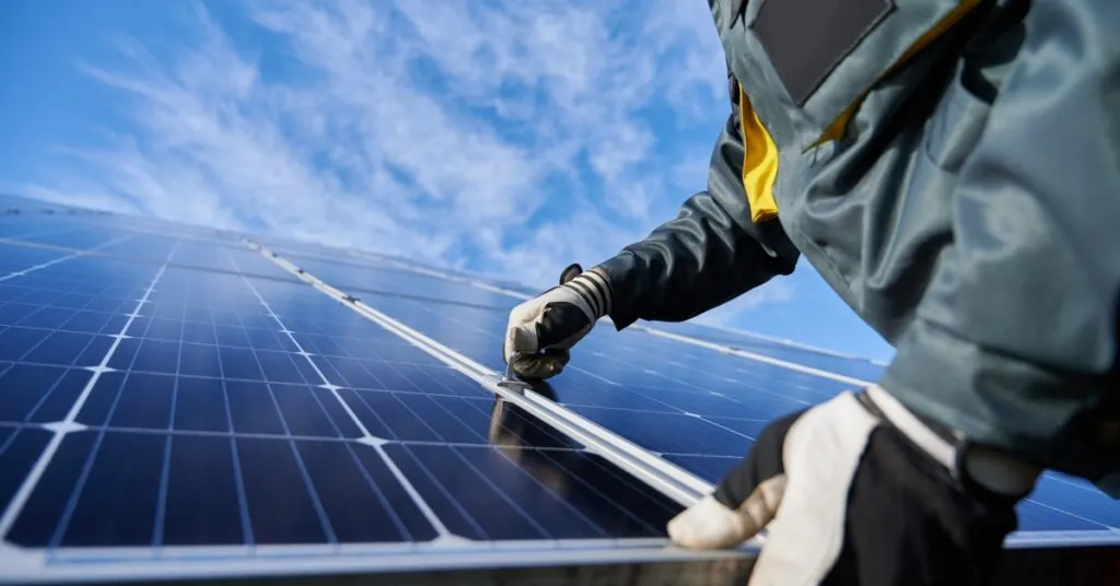 Worker wearing protective gloves and a heavy-duty jacket installing a large solar panel on a sunny day.