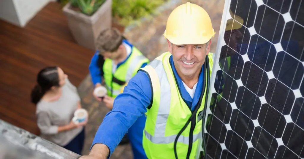 How PV Panel Installation Has Evolved With Modern Technology 2 A worker wearing a yellow hard hat and safety vest climbs a ladder and carries a solar panel to the roof of a home.