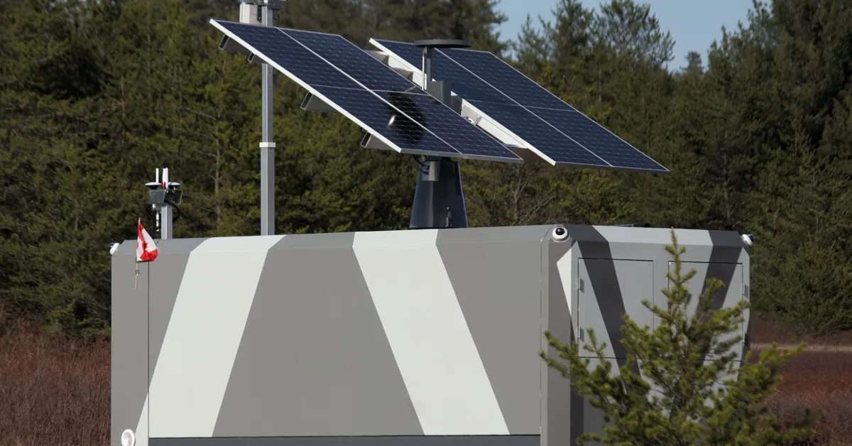 Learn 7 Large grey and white battery with solar panels attached to the top, placed on a plot of land surrounded by trees.