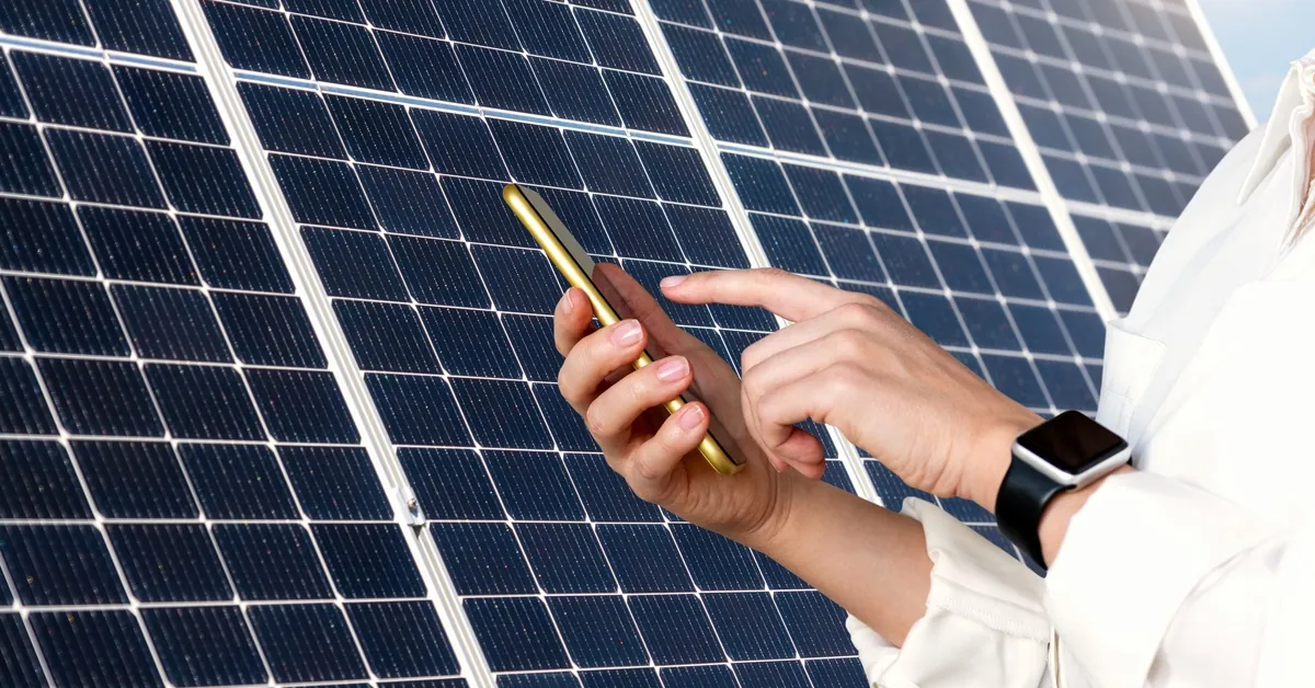 A close-up of the hands of a man holding a smartphone and touching the screen in front of a large solar panel.