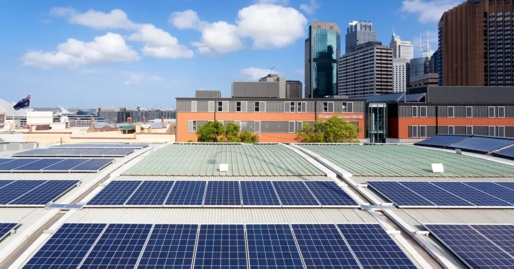 An aerial view of multiple solar panel rows on a large roof with multiple buildings in the background on a sunny day.