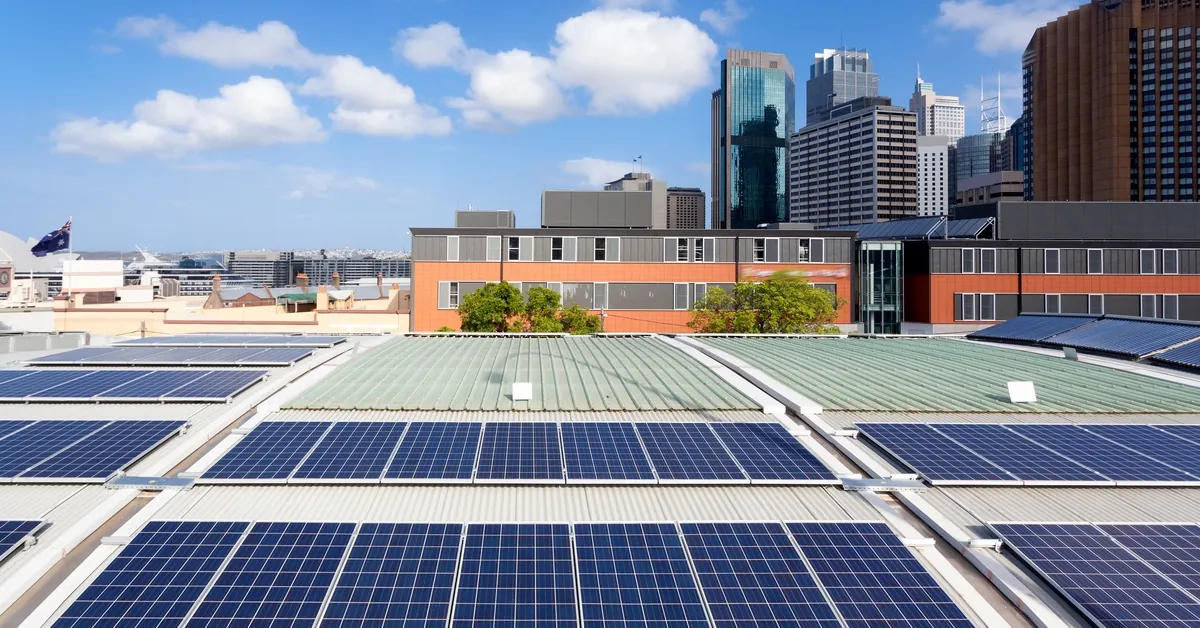 Learn 4 An aerial view of multiple solar panel rows on a large roof with multiple buildings in the background on a sunny day.