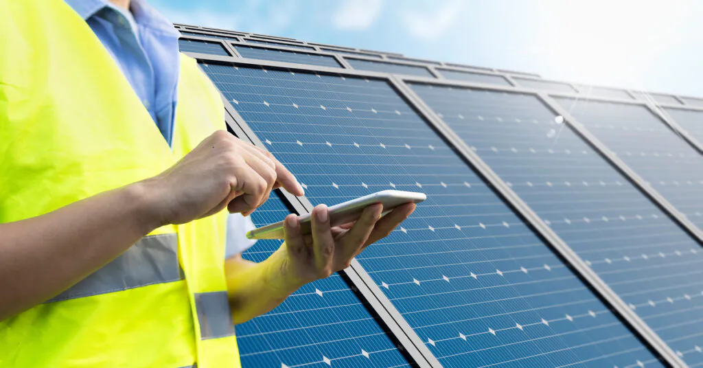 Solar Monitoring Systems Performance Data Access 1 A close-up to the hands of a man wearing a yellow vest, holding a smartphone with solar panels in the background.