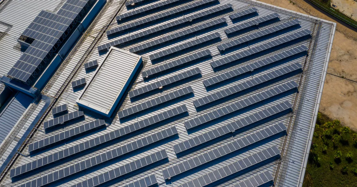 Installing Commercial Solar Panels: Asphalt vs. Metal Roofs 2 An aerial view of a large building with a metal roof with multiple rows of solar panels under bright sunlight.