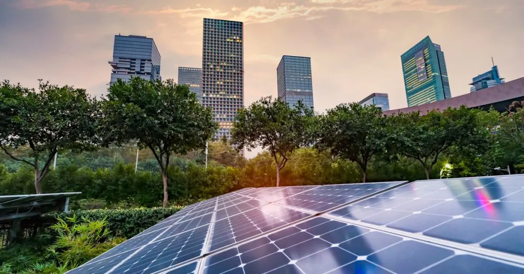 Three-Phase Power and Commercial Solar Integration 2 A close-up of a long solar panel next to an area covered in bushes and trees with multiple buildings in the background.