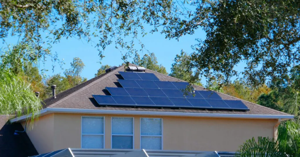 A two-story home with four rows of solar panels placed in a pyramid shape on top of the roof next to multiple trees.