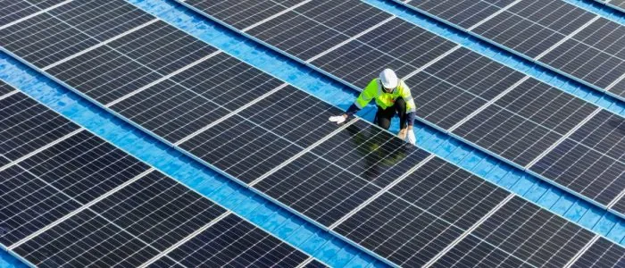 A worker wearing a bright yellow jacket and a white hard hat kneels next to a row of solar panels on a roof.