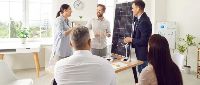 A group of people gathered in a meeting room, talking together. One man is holding a small solar panel on top of a desk.