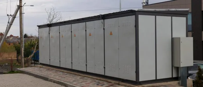 A large white battery on a concrete slab with six pairs of doors, with electrical warning labels placed outdoors.